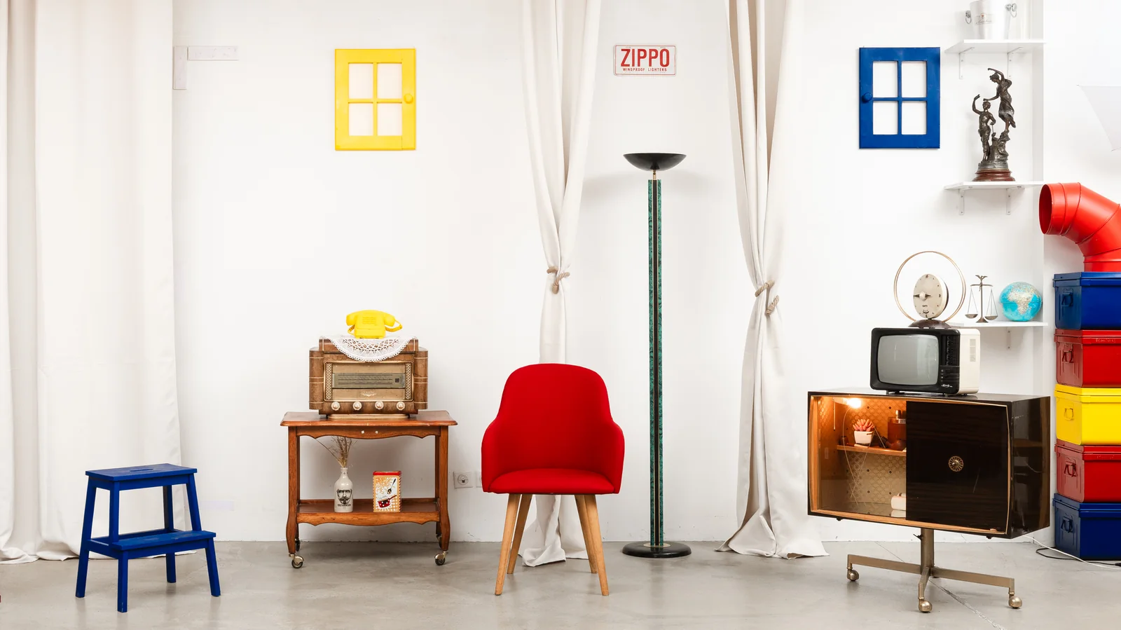 Le Petit Salon rétro vu de face : fenêtres jaune et bleue, plaque Zippo, fauteuil rouge, torchère, meuble TV, desserte radio et escabeau bleu